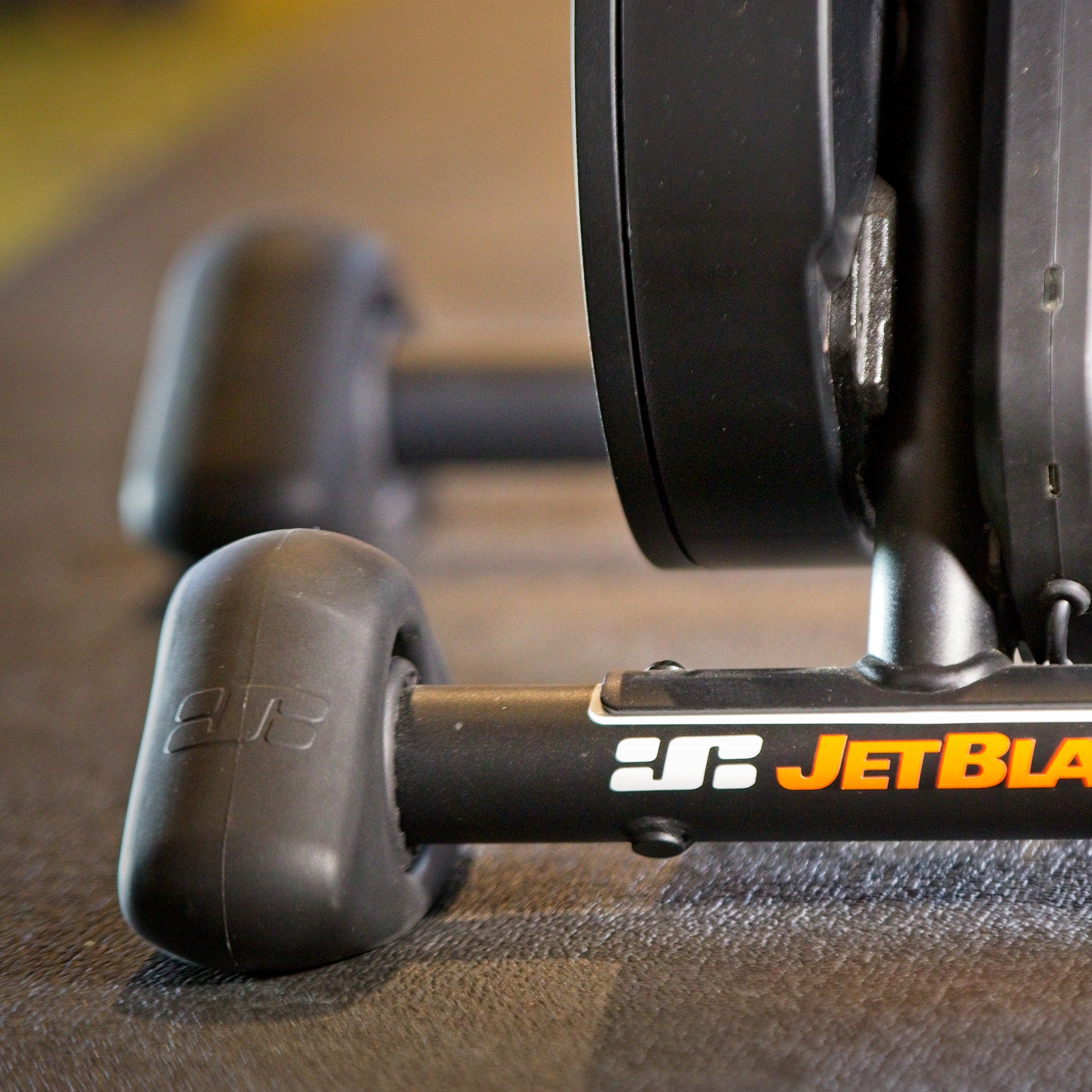 Close-up of a JetBlack indoor cycling trainer with black wheels and a black frame, featuring the brand name in orange on the central bar, set on a gym floor.