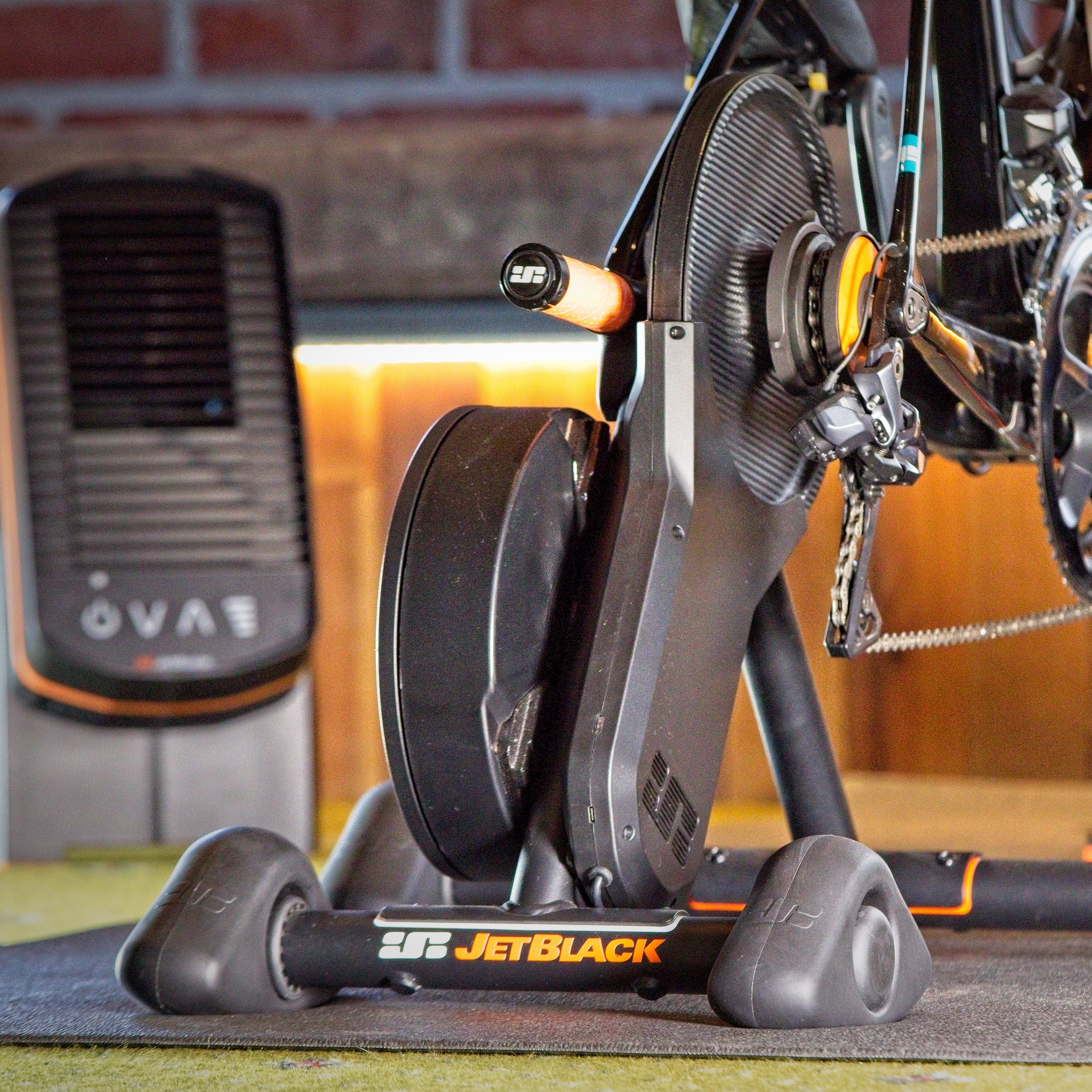 A close-up of a cyclist's lower legs and pedal in motion while using a JetBlack indoor cycling trainer with a flywheel, set in a gym with a brick wall and warm wood paneling in the background.
