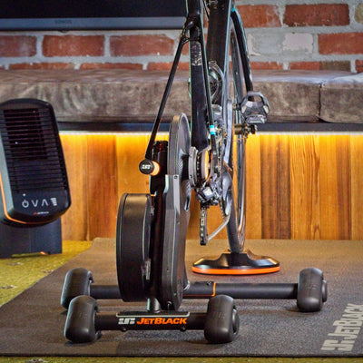 A road bike is mounted on a black indoor trainer setup with a fan and a mat on a gym floor, with a brick and wood wall in the background.
